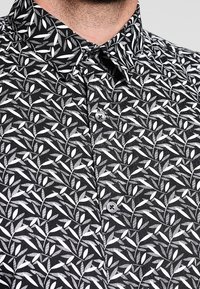Czarna koszula z białym wzorem liści, z kołnierzykiem zapinanym na guziki i plisą na guziki z przodu. Materiał ma gładką fakturę.