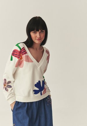 Žena s tmavými vlasy, oblečená v bílém svetru s výstřihem do V a barevnými květinovými vzory a volných modrých kalhotách s malými vyšívanými květy.