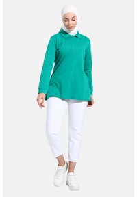 Polo shirt turchese a maniche lunghe con colletto, abbinato a pantaloni bianchi alla caviglia e sneakers bianche, indossato da una persona in hijab.