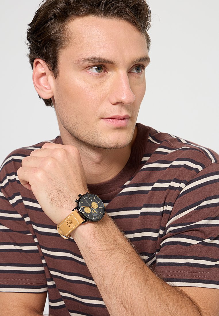 Czarny analogowy zegarek z złotymi akcentami i brązowym skórzanym paskiem, noszony na nadgarstku. Paski brązowa koszulka noszona pod spodem.