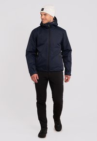Marineblauwe waterdichte jas met capuchon, voorzien van gele ritsaccenten en twee zijzakken, gecombineerd met zwarte slim-fit broek en laarzen.