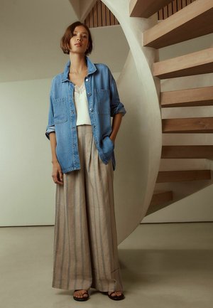 Frau steht drinnen neben einer geschwungenen Holztreppe und trägt eine blaue Jeansjacke, ein weißes Spitzenoberteil, weit geschnittene beige Hosen mit Streifen und schwarze Sandalen.