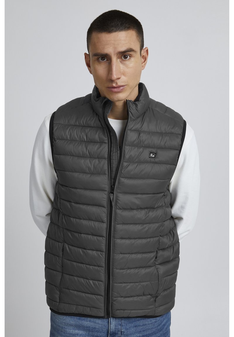 Chaleco puffer gris con cuello alto, diseño acolchado horizontal, cremallera frontal y bolsillos laterales. Llevado sobre una camisa de manga larga blanca.