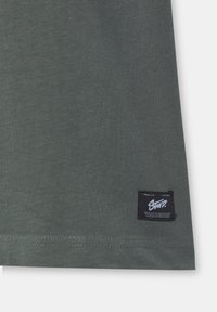 T-shirt di cotone verde scuro con orlo diritto, caratterizzato da un'etichetta nera con testo bianco sul lato sinistro in basso. Texture liscia e design semplice.