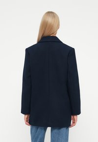 Donna con lunghi capelli biondi, che indossa un cappotto sartoriale blu navy e jeans blu, in piedi di spalle su uno sfondo bianco.