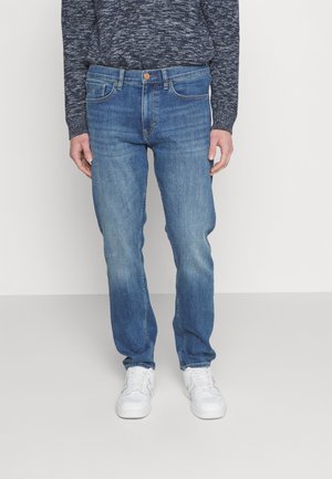 Person iført blå udvaskede jeans med lige ben, en mørkegrå mønstret sweater og hvide sneakers, stående mod en ensfarvet lys baggrund.