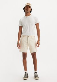 Shorts de denim claro con efecto desgastado y bordes deshilachados, combinados con una camiseta blanca sencilla y zapatillas negras, accessorios con una gorra beige.