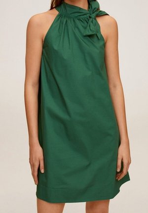 Robe verte sans manches jusqu'aux genoux avec un col montant plissé noué sur le côté gauche, portée par une personne debout.