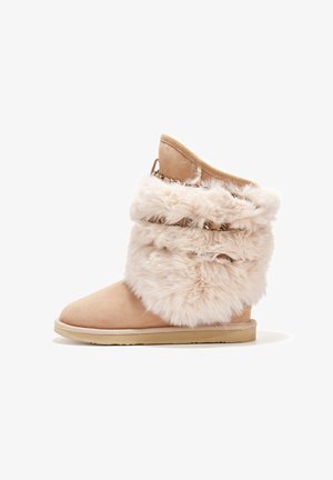 Bota de ante beige con puño de pelo sintético esponjoso, puntera redondeada, suela plana y herrajes decorativos en el puño. Textura suave en todo.