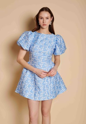 Vrouw draagt een korte blauwe bloemenjurk met pofmouwen, staat met handen gevouwen voor zich tegen een beige achtergrond.