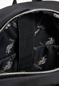 Černý nylonový vnitřek batohu s hladkou texturou, obsahující opakující se světlý vzor s logem U.S. Polo Assn. a látkový pásek.