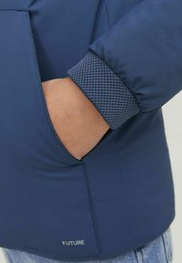 Mörkblå jacka med en slät yta, texturerad meshmanschett och en sidoficka. Har ordet "FUTURE" nära fållen.