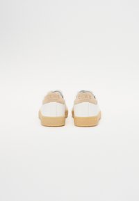 White sneakers with beige accents, smooth leather upper, textured heel tab, and gum rubber sole. Branding visible on heel.
