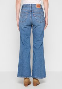 Personne portant un jean Levi's bleu à jambes larges avec poches arrière visibles, debout les mains détendues le long du corps, portant des bottines beige à talons.