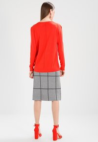 Femme debout, tournée de dos, portant un haut à manches longues rouge vif, une jupe grise atteignant les genoux avec un motif en grille noire, et des sandales à talons rouges.