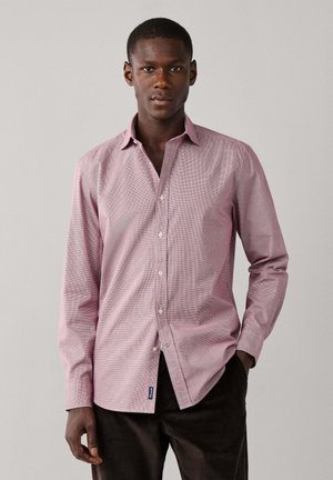 Hombre con camisa rosa de manga larga abotonada y pantalones oscuros, de pie con una mano en el bolsillo contra un fondo liso.