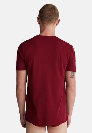 Man met kort haar in een effen bordeauxrood T-shirt met korte mouwen, gezien van achteren tegen een effen witte achtergrond.
