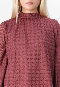 Blonde vrouw in een mauve kant blouse met hoge kraag, gemaakt van ingewikkeld geometrisch patroon stof.