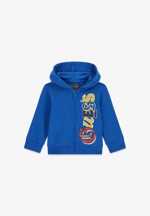 Sweat à capuche bleu zippé avec logo coloré "GUESS" imprimé verticalement sur le côté droit et poignets et bas côtelés.