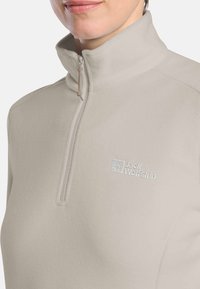 Ingrandimento di un pullover in fleece beige chiaro con colletto alto con zip e logo Jack Wolfskin sul petto, indossato da una persona.