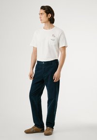 Camiseta blanca de manga corta con detalle de bolsillo, combinada con pantalones de pana azul marino, complementada con zapatillas marrones con acentos azules.
