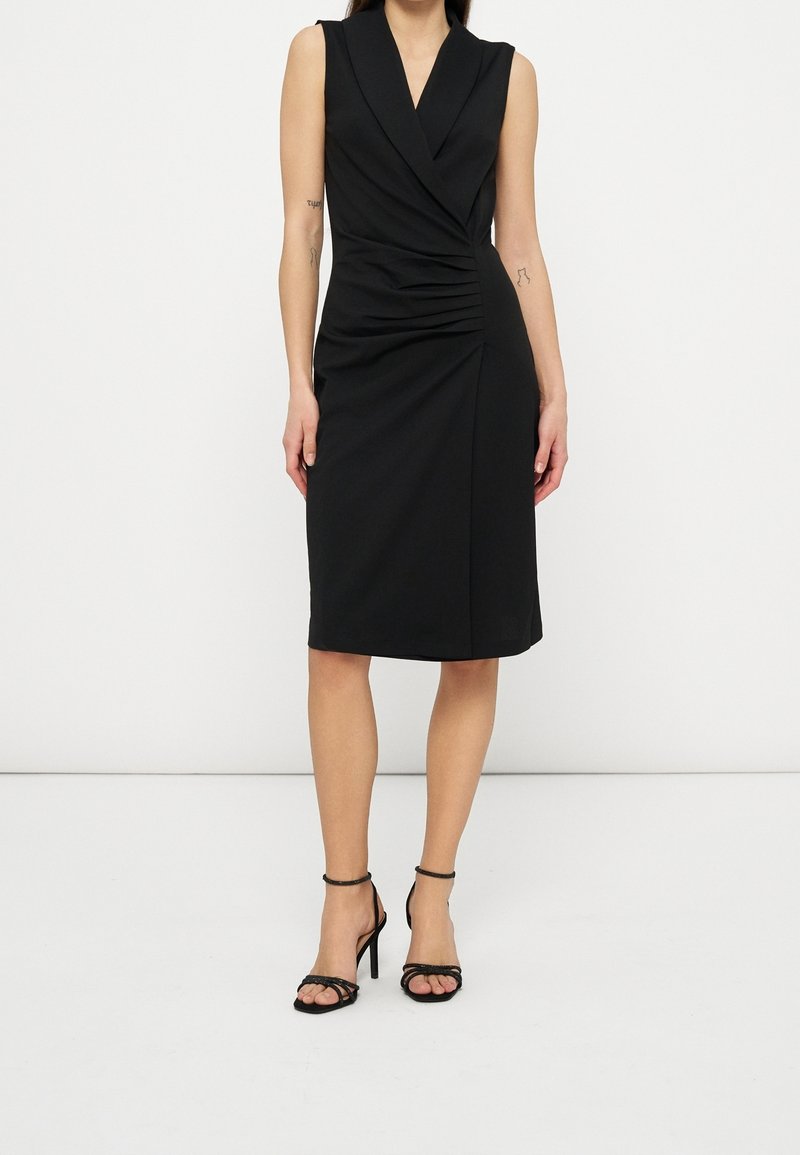 Robe noire sans manches longueur genou avec devant style portefeuille et détails froncés, portée avec des sandales noires à talons hauts et brides à la cheville.