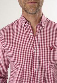 Camisa de cuadros pequeña en rojo y blanco con cuello puntiagudo y dos cierres de botón. Presenta un pequeño logo en el lado izquierdo del pecho.