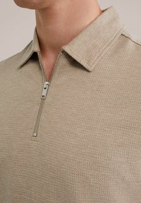 Polo de cuello con cremallera en color beige, confeccionado con un tejido de punto texturizado y con un cuello puntiagudo. Presenta una cremallera metálica plateada y un diseño ajustado.