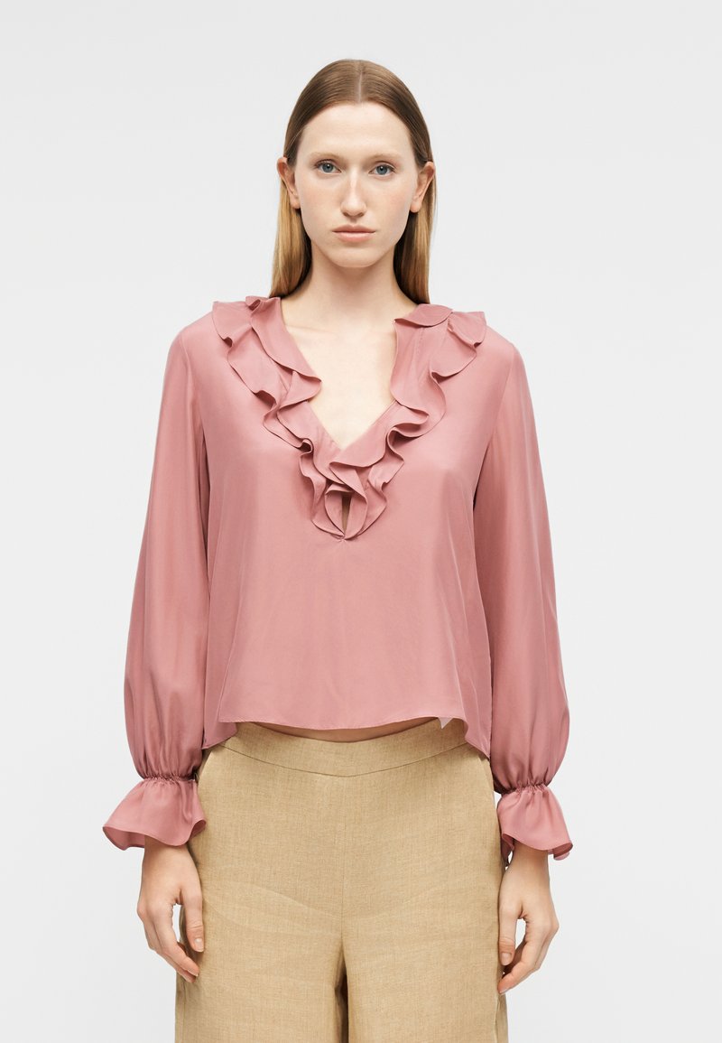 Femme aux cheveux raides portant un chemisier rose poudré à volants avec un col en V et un pantalon beige taille haute à jambes larges, debout devant un fond uni.