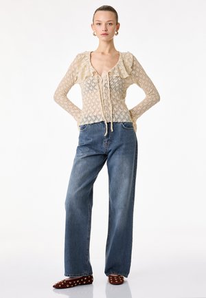 Mujer de pie vistiendo una blusa beige de encaje con volantes, vaqueros azules holgados y bailarinas rojas con tachuelas sobre un fondo blanco.