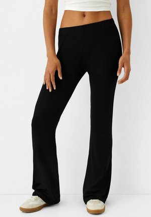 Kvinde iført sorte rillede vidde bukser, hvid cropped tank top og hvide sneakers med brune såler mod en ensfarvet hvid baggrund.