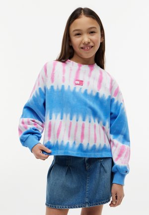 Młoda dziewczyna uśmiecha się, nosi różowo-niebieską bluzę tie-dye i niebieską dżinsową spódnicę, stoi na jednolitym białym tle.
