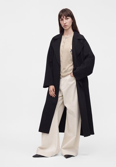 Femme aux longs cheveux bruns portant un long manteau noir, un cardigan beige, un pantalon blanc large et des chaussures noires pointues sur un fond uni.