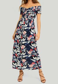 Robe maxi bleu marine à épaules dégagées avec un motif floral en rouge, rose et blanc, dotée d'un corsage smocké et d'une jupe évasée. Sandales à brides marron.