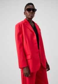 Red tailored blazer made of smooth fabric, featuring a notch lapel, two buttons, and side pockets. Worn over a black top.