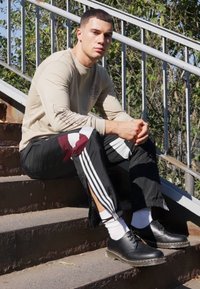 Camisa beige de manga larga con logo, pantalones deportivos negros con detalles en burdeos y blanco, calcetines blancos y zapatillas negras. Sentado en escaleras al aire libre.