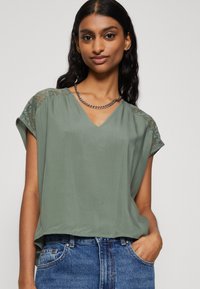 Grønn, lett bluse med V-hals og blonde detaljer på skuldrene. Korte ermer og avslappet passform, kombinert med blå denimjeans.