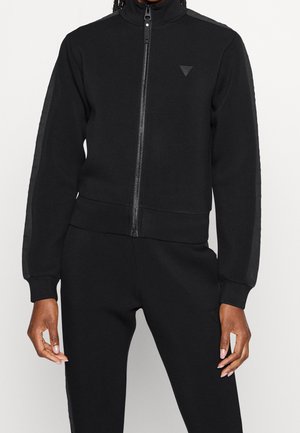 Person wearing a black zip-up jacket with subtle side stripes and matching black pants, standing against a plain background.