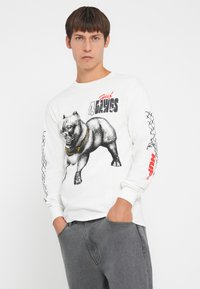 Witte lange mouwen t-shirt met een zwarte afbeelding van een hond, met rode "4 the Days" tekst en een zwart kettingpatroon op de mouwen.