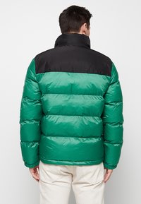 Grüne Puffjacke mit schwarzem Oberteil, ausgestattet mit einem gesteppten Design und hohem Kragen. Das Material wirkt glänzend und glatt.