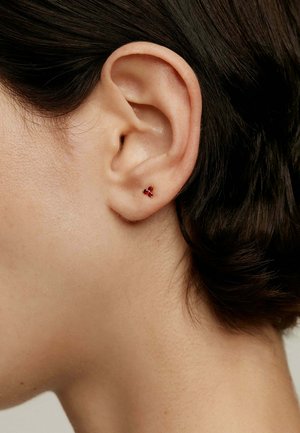 Pendientes de botón de gema roja en un diseño de racimo triangular, montados en una base metálica, exhibidos en un lóbulo de la oreja contra una textura de piel suave.