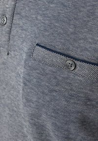Nahaufnahme von einer blauen, strukturierten Stoffhemd-Tasche mit einem kleinen Knopf, der "CASAMODA" beschriftet ist, und einem kontrastierenden dunklen Besatz.