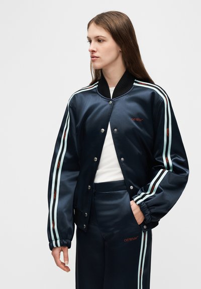 Γυναίκα που φοράει σατέν μπλε-ναυτικό σακάκι και ταιριαστό παντελόνι με ανοιχτό μπλε ρίγες και το λογότυπο "Off-White", κοιτάζοντας προς τα αριστερά.