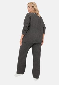 Blondynka w luźnym, ciemnoszarym swetrze i pasujących szerokich spodniach stoi, patrząc w bok na białym tle.