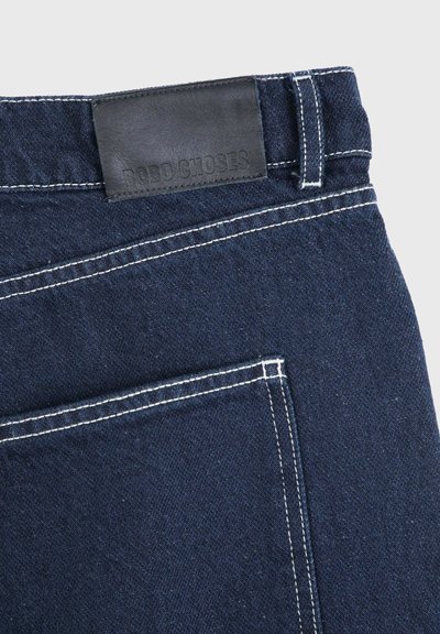 Jean en denim bleu foncé à l'arrière avec des surpiqûres blanches et un patch de marque en cuir noir près de la ceinture au-dessus de la poche.
