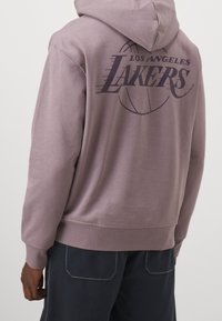 Felpa viola in morbido tessuto con la scritta "LOS ANGELES LAKERS" e grafica di un pallone da basket sul retro. Tasca a marsupio frontale.