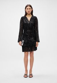 Vestido preto com paetês, decote em V, mangas longas tipo sino e cintura franzida. Apresenta uma bainha em camadas com babados e é combinado com sandálias pretas.