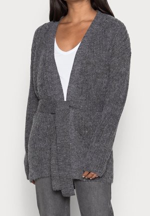 Femme portant un cardigan tricoté gris foncé avec ceinture nouée à la taille, par-dessus un haut blanc et un jean gris, sur un fond uni.