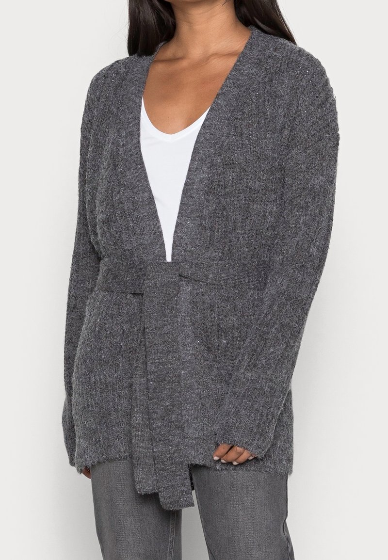 Femme portant un cardigan tricoté gris foncé avec ceinture nouée à la taille, par-dessus un haut blanc et un jean gris, sur un fond uni.