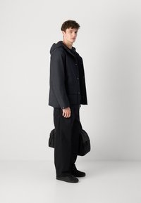 Veste imperméable noire avec capuche, boutons sur le devant et poches latérales, associée à un pantalon noir et un sac noir, se tenant devant un fond neutre.